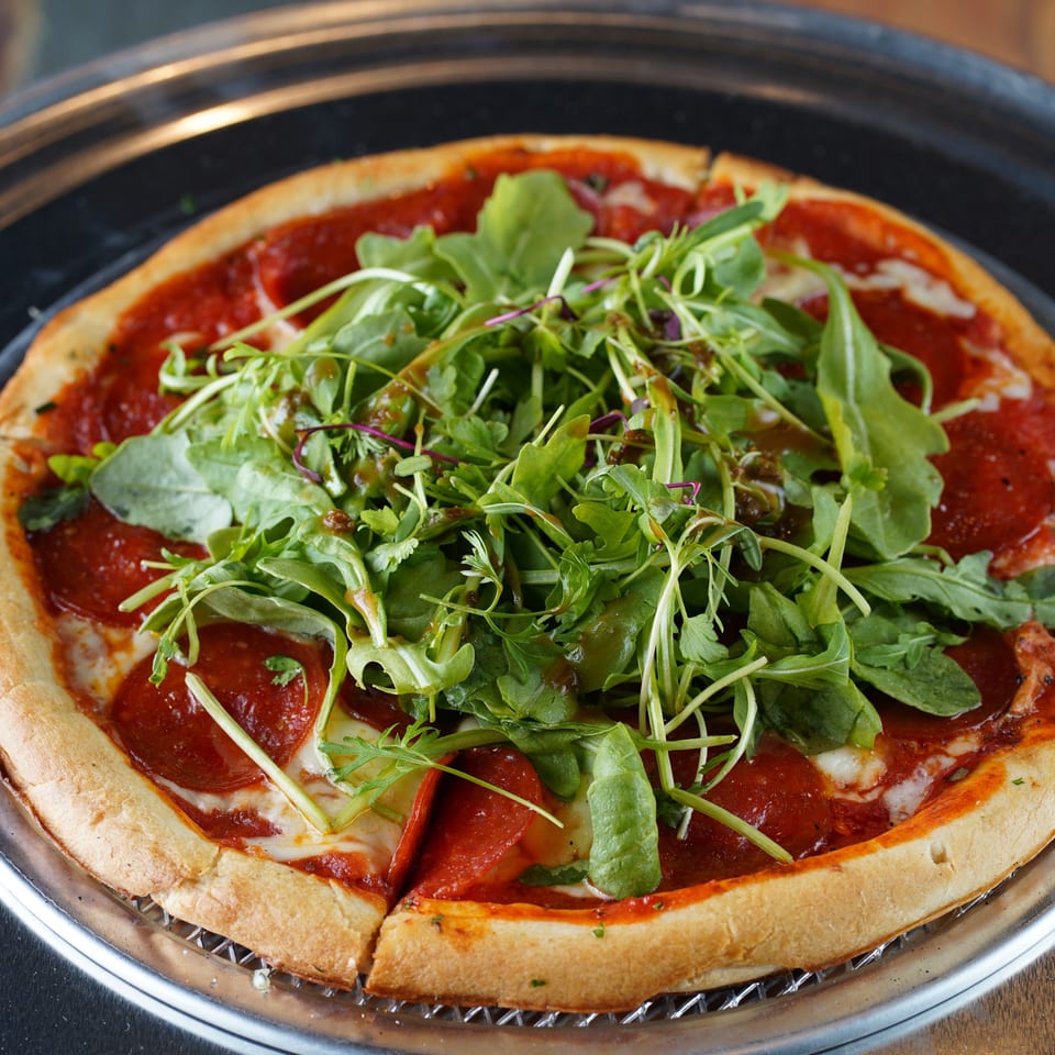 Pepperoni pizza topped with fresh arugula and balsamic glaze on a metal tray.