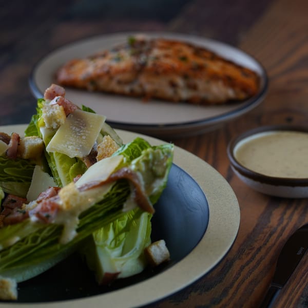 Caesar salad with croutons and parmesan, a side of dressing, and a salmon fillet on a wooden table.
