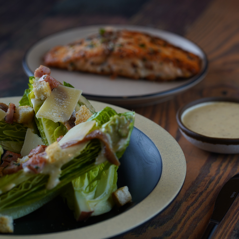 Caesar salad with croutons and parmesan, a side of dressing, and a salmon fillet on a wooden table.