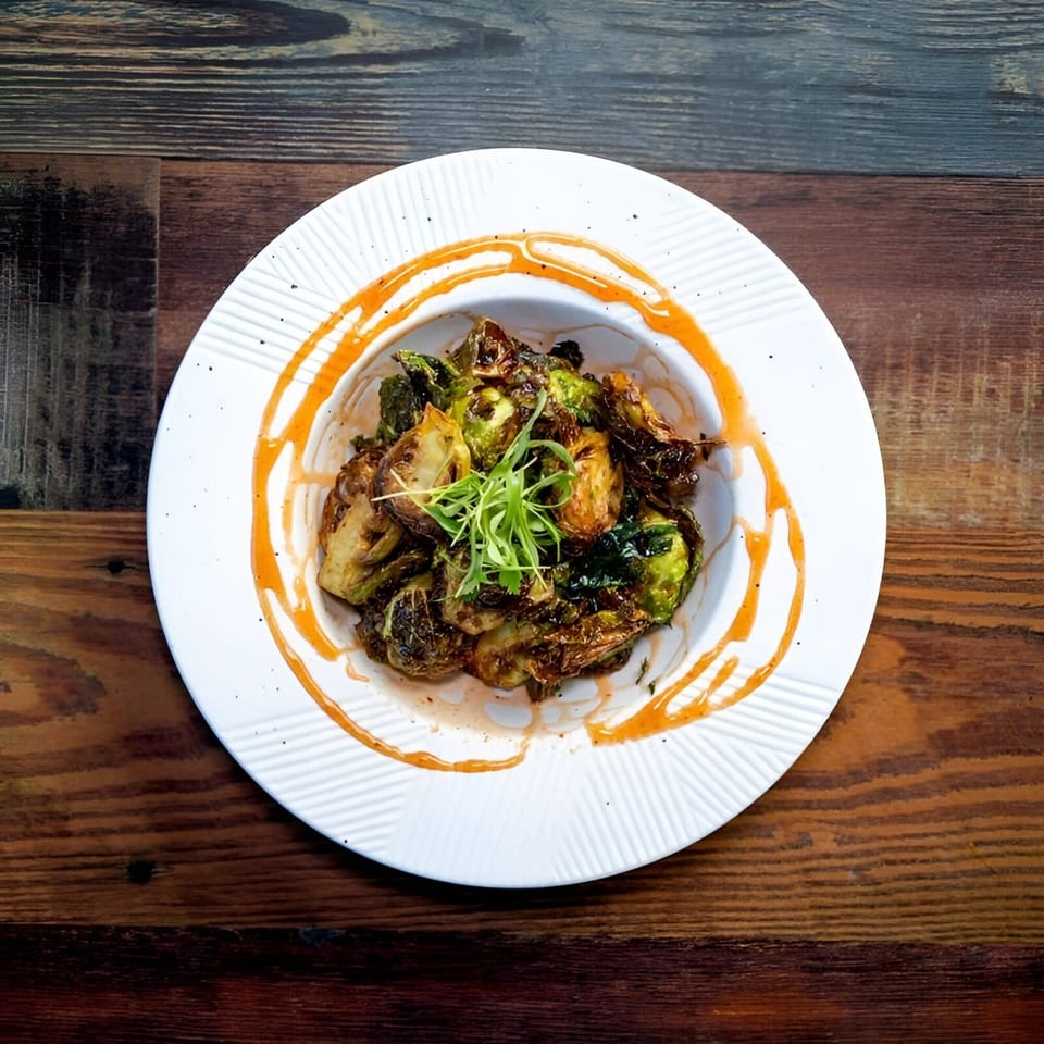 A white bowl of crispy Brussels sprouts with a drizzle of orange sauce and green garnish.