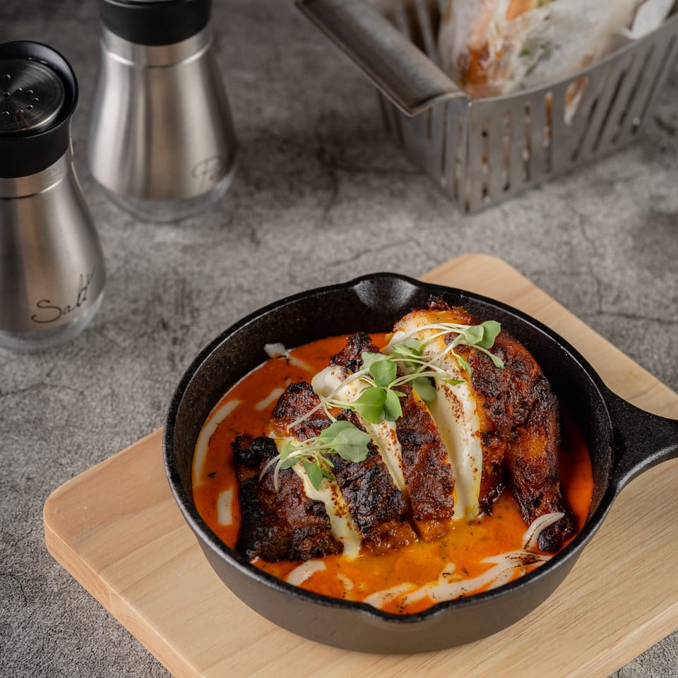 Grilled chicken breast in a skillet with red sauce, white drizzle, and microgreens on a wooden board.