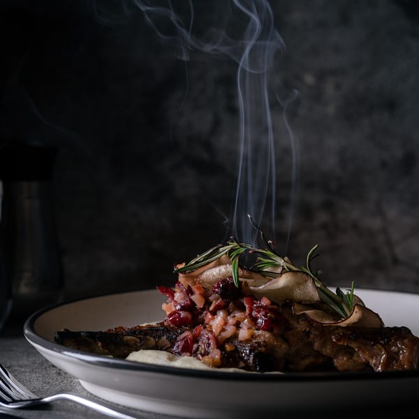 Steaming grilled pork chop with cranberry relish, rosemary, and potato puree on a dark background.
