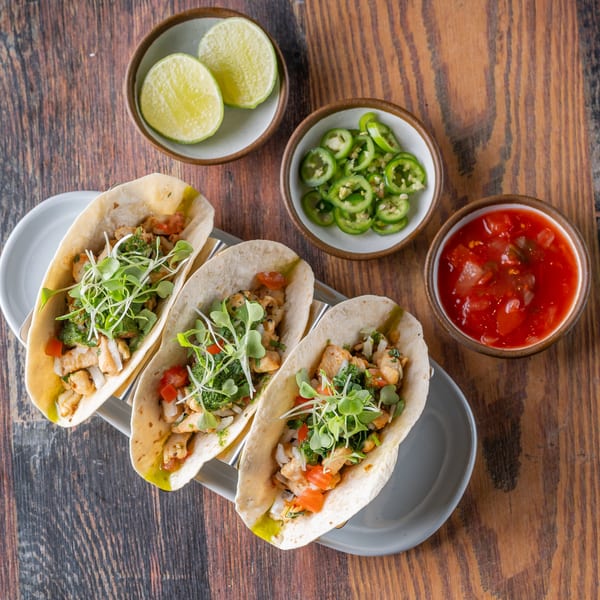Three chicken tacos with microgreens, salsa, sliced jalapeños, and lime wedges on a wooden table.