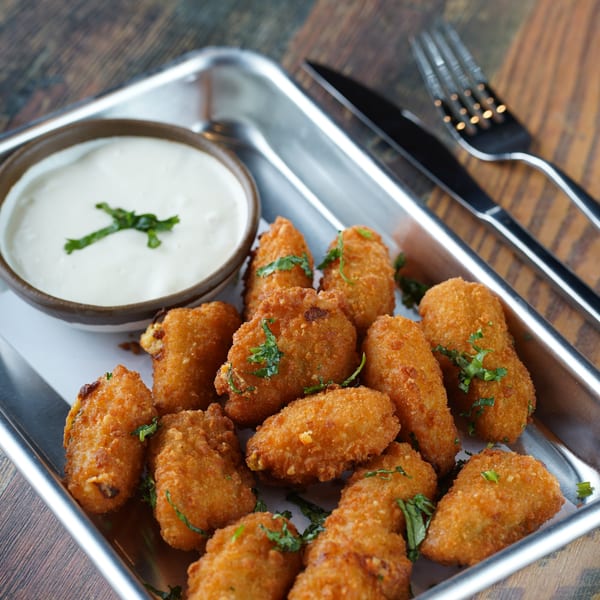 Crispy fried appetizers with a side of creamy white dipping sauce, garnished with herbs on a metal tray.
