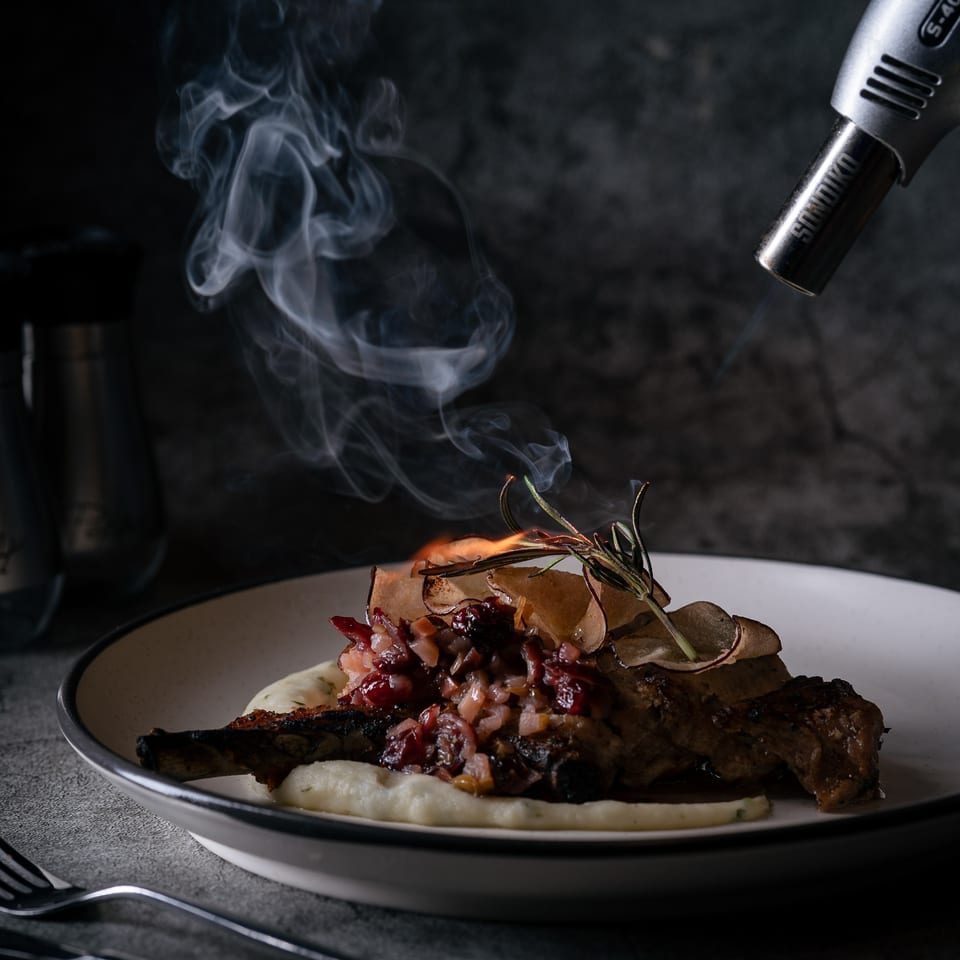 A chef's torch caramelizing a pork chop with mashed potatoes and a red relish, creating smoke.